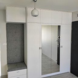 MEBLEKO - Biała zabudowa przedpokoju z siedziskiem obitym szarą tkaniną, lustrzane drzwi szafy, oświetlenie sufitowe w formie białej kuli.