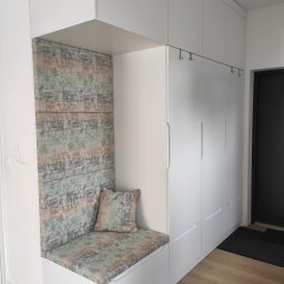 MEBLEKO - Biała zabudowa przedpokoju z siedziskiem i tapicerowanym panelem ściennym w odcieniach szarości, zieleni i beżu, uzupełniona poduszką w dopasowanym wzorze, z minimalistycznymi wieszakami na ubrania.