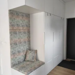 Biała zabudowa przedpokoju z siedziskiem i tapicerowanym panelem ściennym w odcieniach szarości, zieleni i beżu, uzupełniona poduszką w dopasowanym wzorze, z minimalistycznymi wieszakami na ubrania.