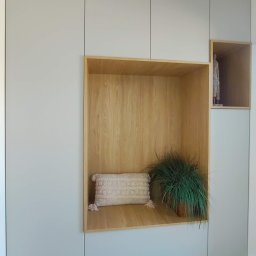 MEBLEKO - Szara zabudowa meblowa z drewnianą wnęką, w której znajduje się poduszka z frędzlami i doniczkowa roślina; dodatkowo mała drewniana wnęka z figurką.