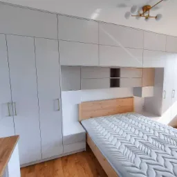 Biała zabudowa szaf w sypialni z drewnianym łóżkiem i szafkami nocnymi wkomponowanymi w zabudowę, widoczne złote uchwyty szafek i lampa sufitowa.