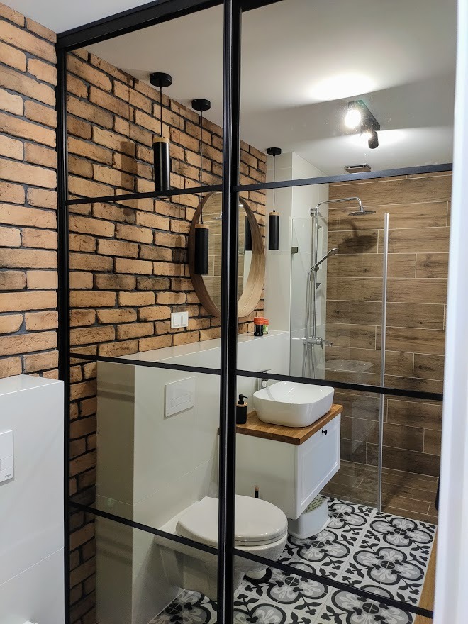 Wnętrze łazienki z kabiną prysznicową oddzieloną czarnymi, przeszklonymi ramami, ceglana ściana, okrągłe lustro i wzorzysta podłoga.