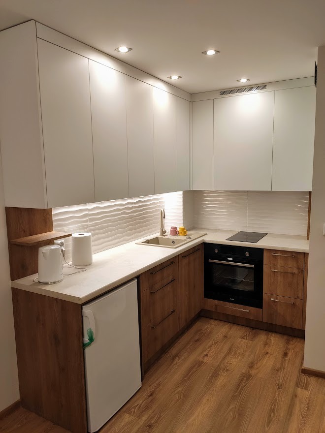 Nowoczesna, narożna kuchnia z białymi górnymi szafkami i drewnopodobnymi dolnymi, z blatem w kolorze jasnego kamienia, falistymi białymi płytkami na ścianie, małą lodówką i płytą indukcyjną.