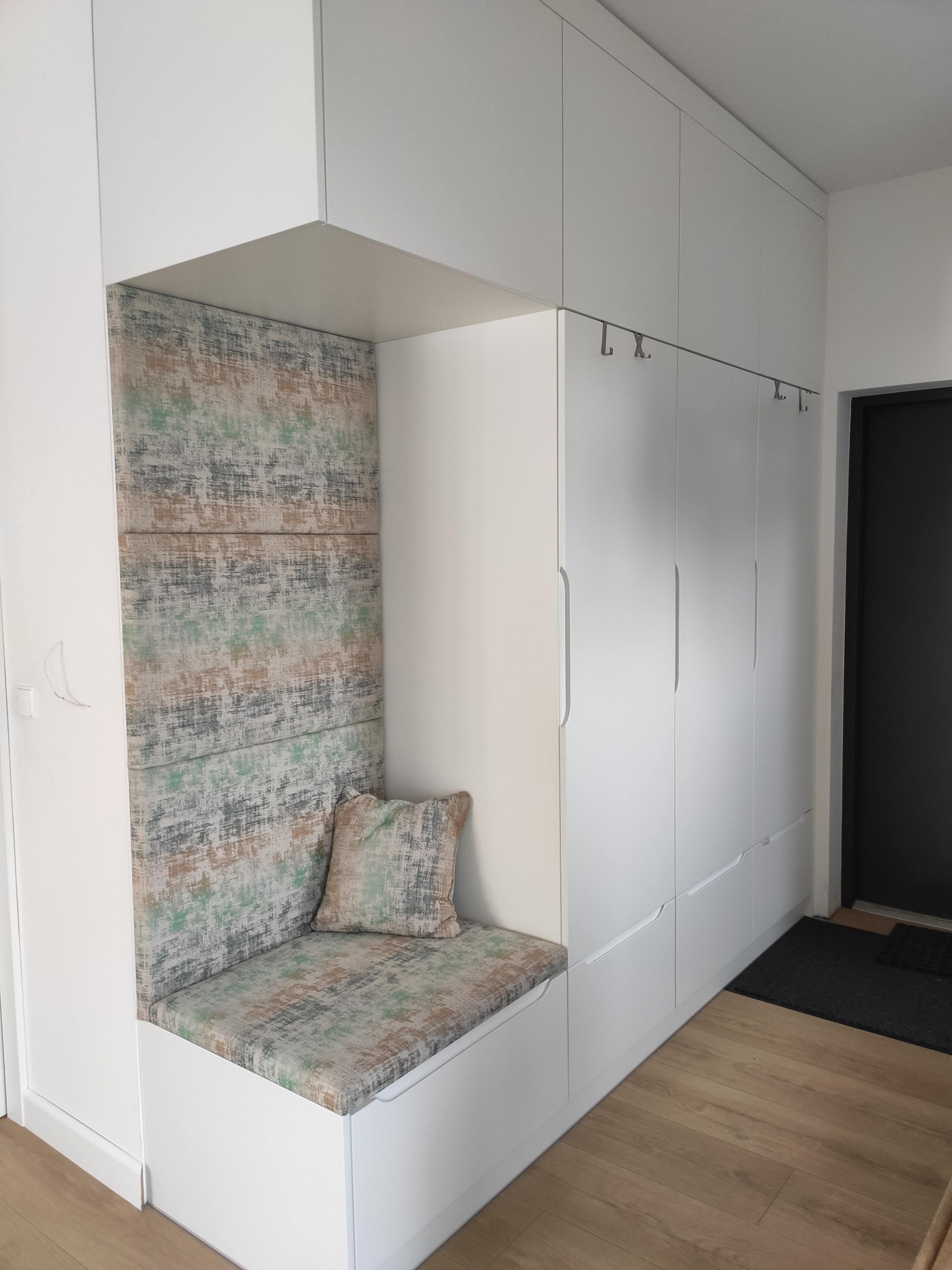 Biała zabudowa przedpokoju z siedziskiem i tapicerowanym panelem ściennym w odcieniach szarości, zieleni i beżu, uzupełniona poduszką w dopasowanym wzorze, z minimalistycznymi wieszakami na ubrania.