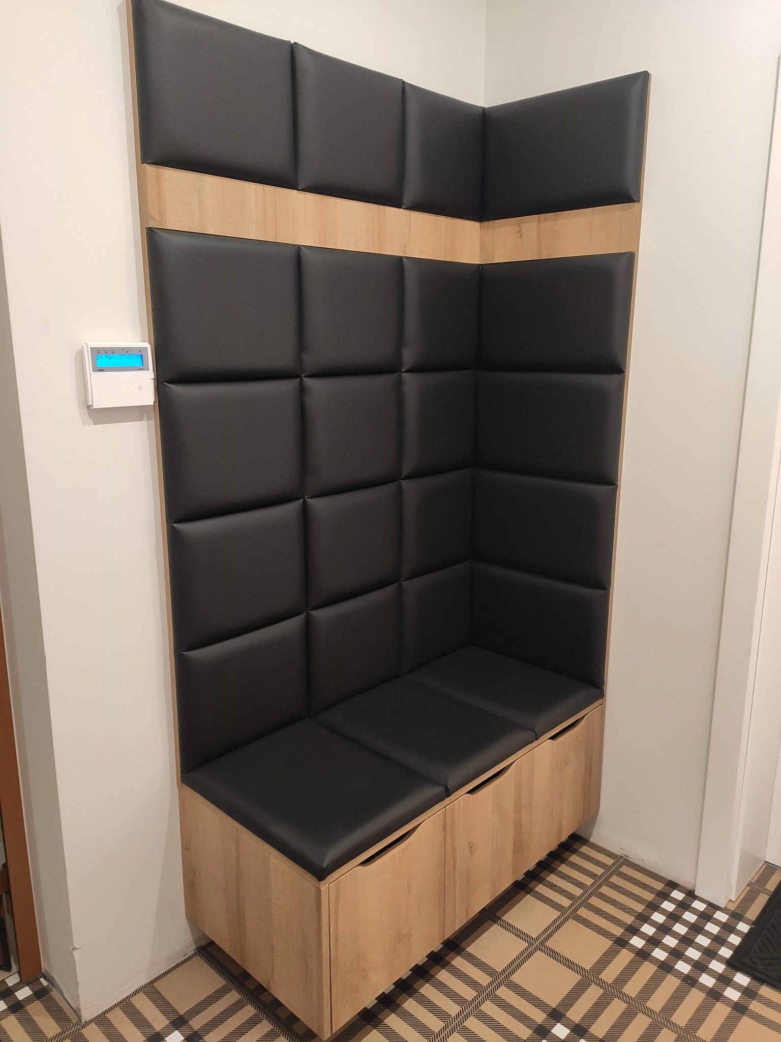 Wnęka z siedziskiem i tapicerowanym panelem ściennym w czarnej skórze, wykończona drewnem o jasnym odcieniu, na podłodze wzorzyste płytki.