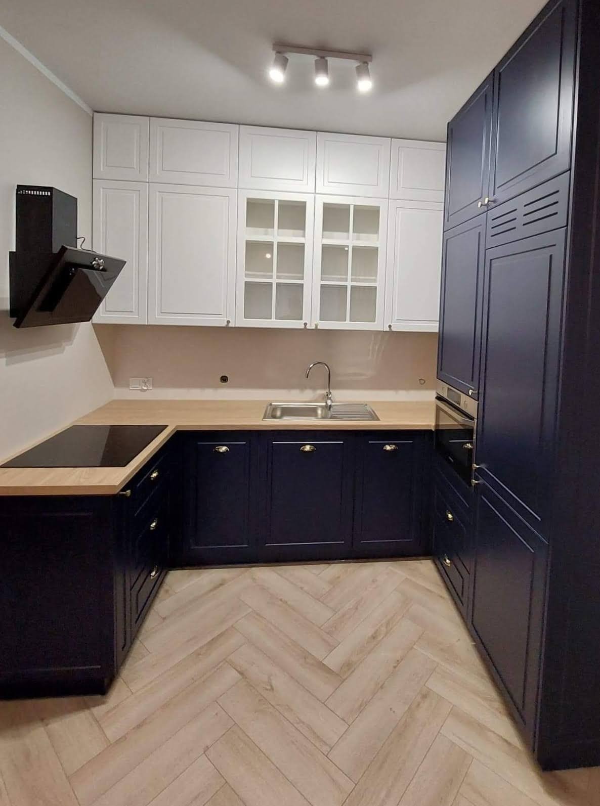 Nowoczesna kuchnia w odcieniach bieli i granatu, z drewnianym blatem i podłogą ułożoną w jodełkę, oświetlona punktowymi lampami sufitowymi.