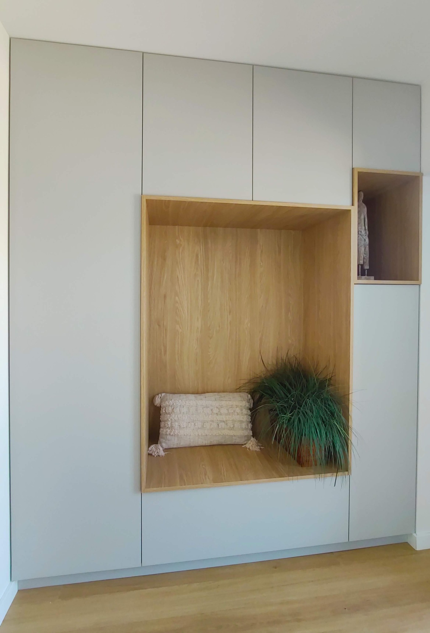 Szara zabudowa meblowa z drewnianą wnęką, w której znajduje się poduszka z frędzlami i doniczkowa roślina; dodatkowo mała drewniana wnęka z figurką.