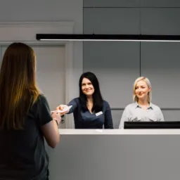 Pacjentka odbiera dokumenty w recepcji centrum medycznego, dwie uśmiechnięte recepcjonistki za ladą, nowoczesne oświetlenie.