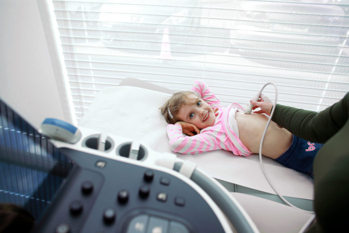 Uśmiechnięta dziewczynka leży na leżance podczas badania USG jamy brzusznej, widoczny aparat USG z licznymi przyciskami i ekranem.