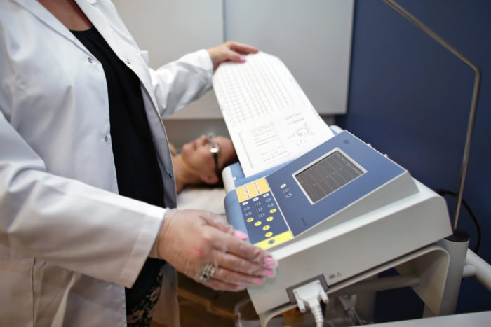 Pacjentka leży na leżance podczas badania EKG; pielęgniarka w białym fartuchu trzyma wydruk z aparatu EKG.