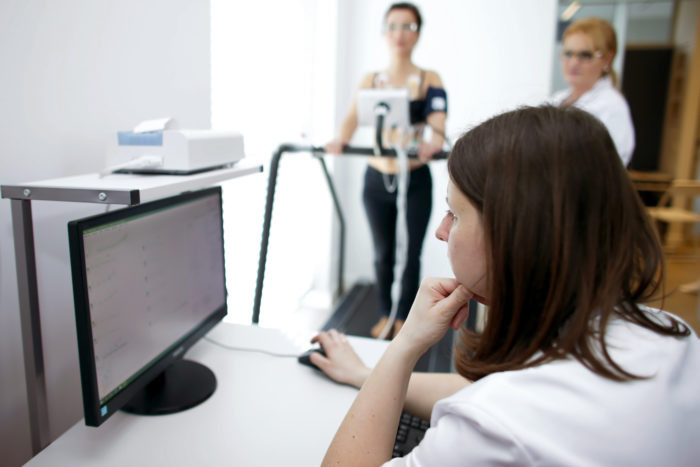 Próba wysiłkowa na bieżni: pacjentka z elektrodami monitorowana przez lekarza przy komputerze, drugi lekarz obserwuje.