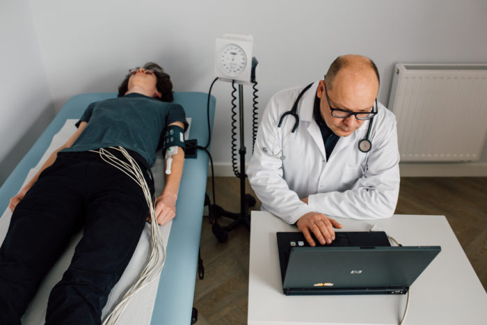 Pacjent leży na leżance z podłączonymi elektrodami EKG i mankietem do pomiaru ciśnienia, lekarz w białym fartuchu i okularach analizuje wyniki na laptopie przy biurku.