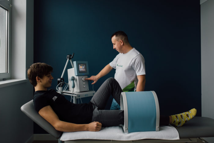 Pacjent leży na leżance podczas zabiegu fizykoterapii z wykorzystaniem aparatu do magnetoterapii, obsługiwanego przez terapeutę w białej koszulce z logo placówki.