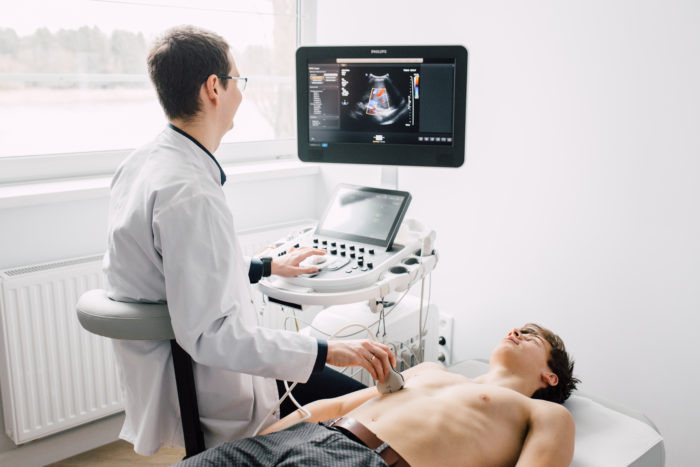Lekarz wykonuje badanie USG serca pacjentowi leżącemu na leżance, widoczny monitor z obrazem serca i aparatura USG.