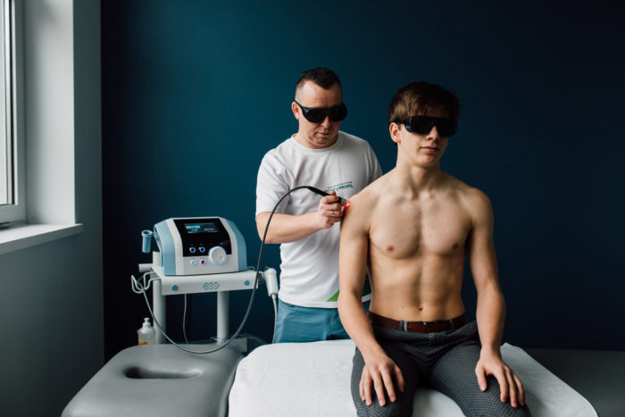 Zabieg laserowy na ramieniu pacjenta w gabinecie w Gdańsku, wykonywany przez terapeutę w okularach ochronnych, widoczny aparat do laseroterapii.