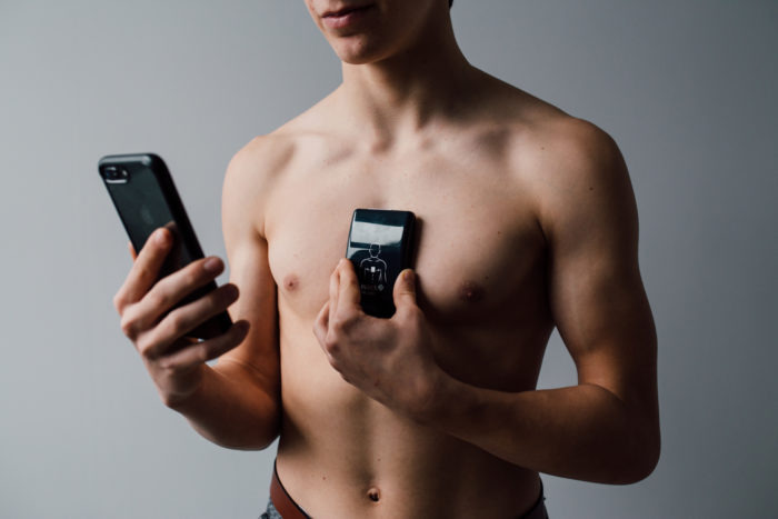 Młody mężczyzna bez koszuli trzyma dwa smartfony: jeden przyłożony do klatki piersiowej z wyświetlonym schematem sylwetki i serca, drugi w drugiej dłoni, prawdopodobnie rejestrujący dane.