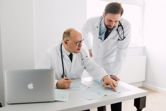 Dwóch kardiologów w białych fartuchach analizuje wyniki EKG przy biurku z laptopem w jasnym gabinecie.