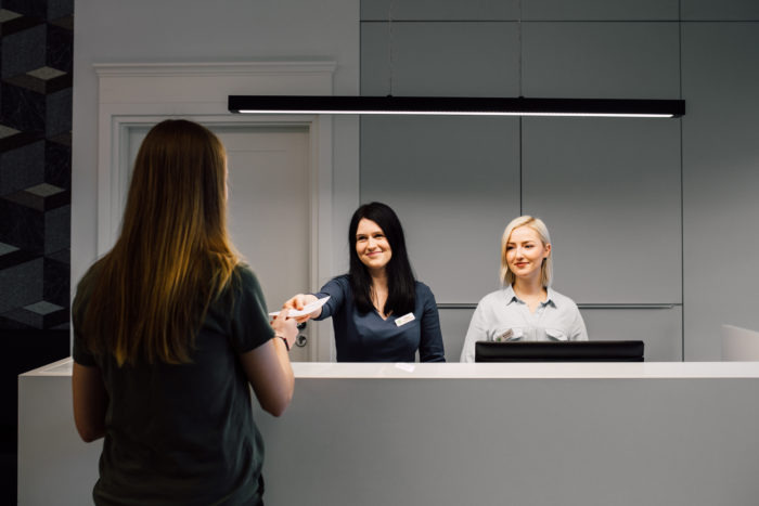 Pacjentka odbiera dokumenty w recepcji centrum medycznego, dwie uśmiechnięte recepcjonistki za ladą, nowoczesne oświetlenie.