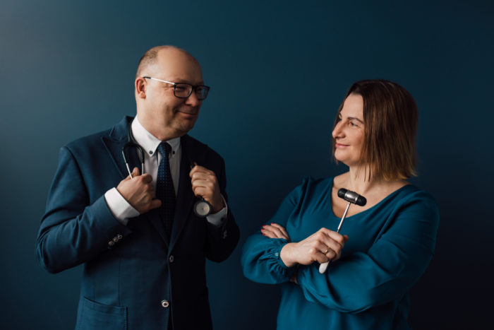 Portret dwojga lekarzy na ciemnym tle: mężczyzna w garniturze ze stetoskopem i zegarkiem, kobieta z młotkiem neurologicznym w ręku, oboje patrzą na siebie z uśmiechem.