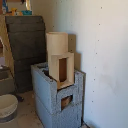 Wnętrze w trakcie budowy z elementami konstrukcyjnymi kominka z szarych bloczków betonowych i beżowych kształtek ceramicznych.