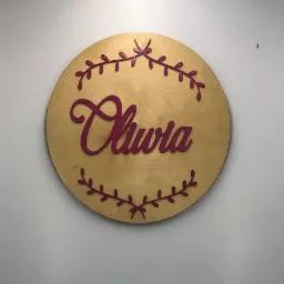 Okrągła, drewniana tabliczka z wyciętym, bordowym napisem 'Olivia' otoczonym dekoracyjnym, bordowym motywem roślinnym, przymocowana do białej ściany.