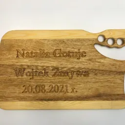 Drewniana deska do krojenia z wygrawerowanym napisem 'Natalia Gotuje, Wojtek Zmywa, 20.08.2021 r.' i uchwytem z czterema otworami na palce.
