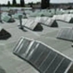 Płaski dach pokryty szarą papą z licznymi świetlikami dachowymi o trójkątnych osłonach, widoczne kominy wentylacyjne i fragmenty zabudowy miejskiej w tle.