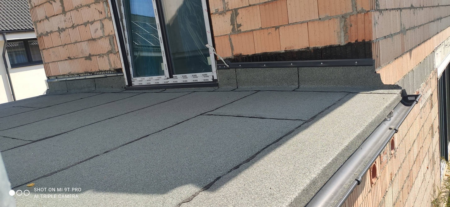 Nowy dach pokryty papą termozgrzewalną, widoczne okno dachowe z folią ochronną, zamontowana rynna w kolorze grafitowym, ściana budynku w trakcie budowy z pustaków ceramicznych.