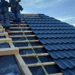 Montaż ciemnografitowej blachodachówki na łatach z widoczną membraną dachową, dwóch dekarzy w uprzężach na dachu, pochmurne niebo w tle.