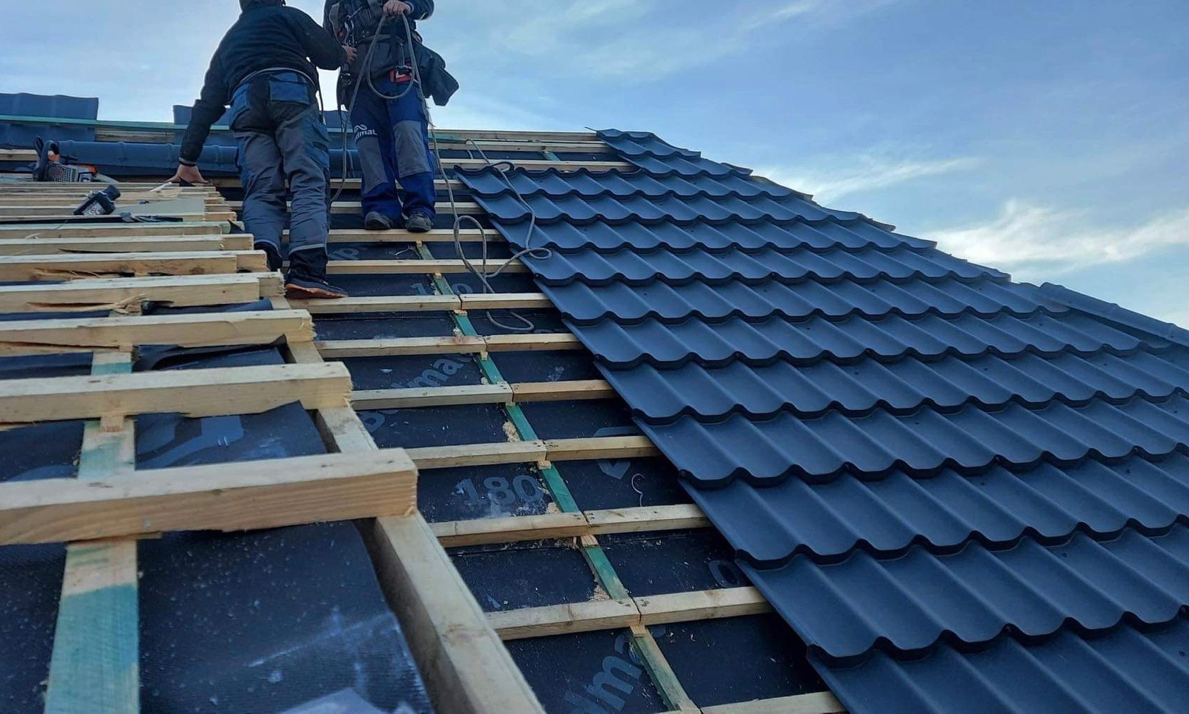 Montaż ciemnografitowej blachodachówki na łatach z widoczną membraną dachową, dwóch dekarzy w uprzężach na dachu, pochmurne niebo w tle.