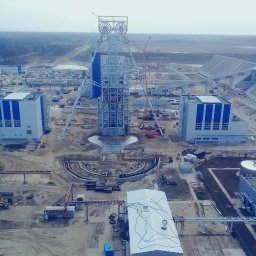 Start - Wysoka wieża wyciągowa z kołami linowymi na szczycie, w trakcie budowy na rozległym placu budowy z budynkami i żurawiami w tle, widok z góry.