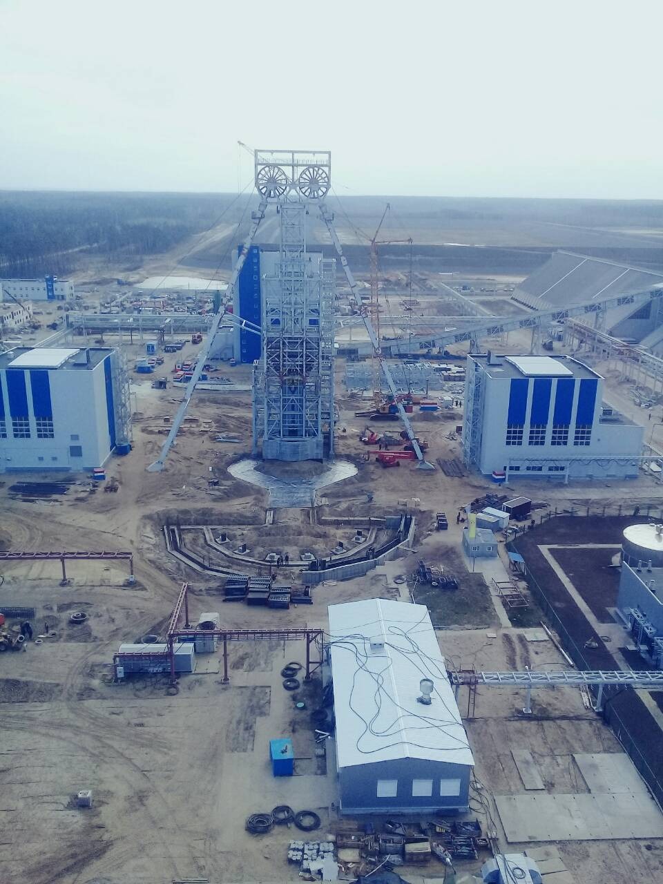 Wysoka wieża wyciągowa z kołami linowymi na szczycie, w trakcie budowy na rozległym placu budowy z budynkami i żurawiami w tle, widok z góry.