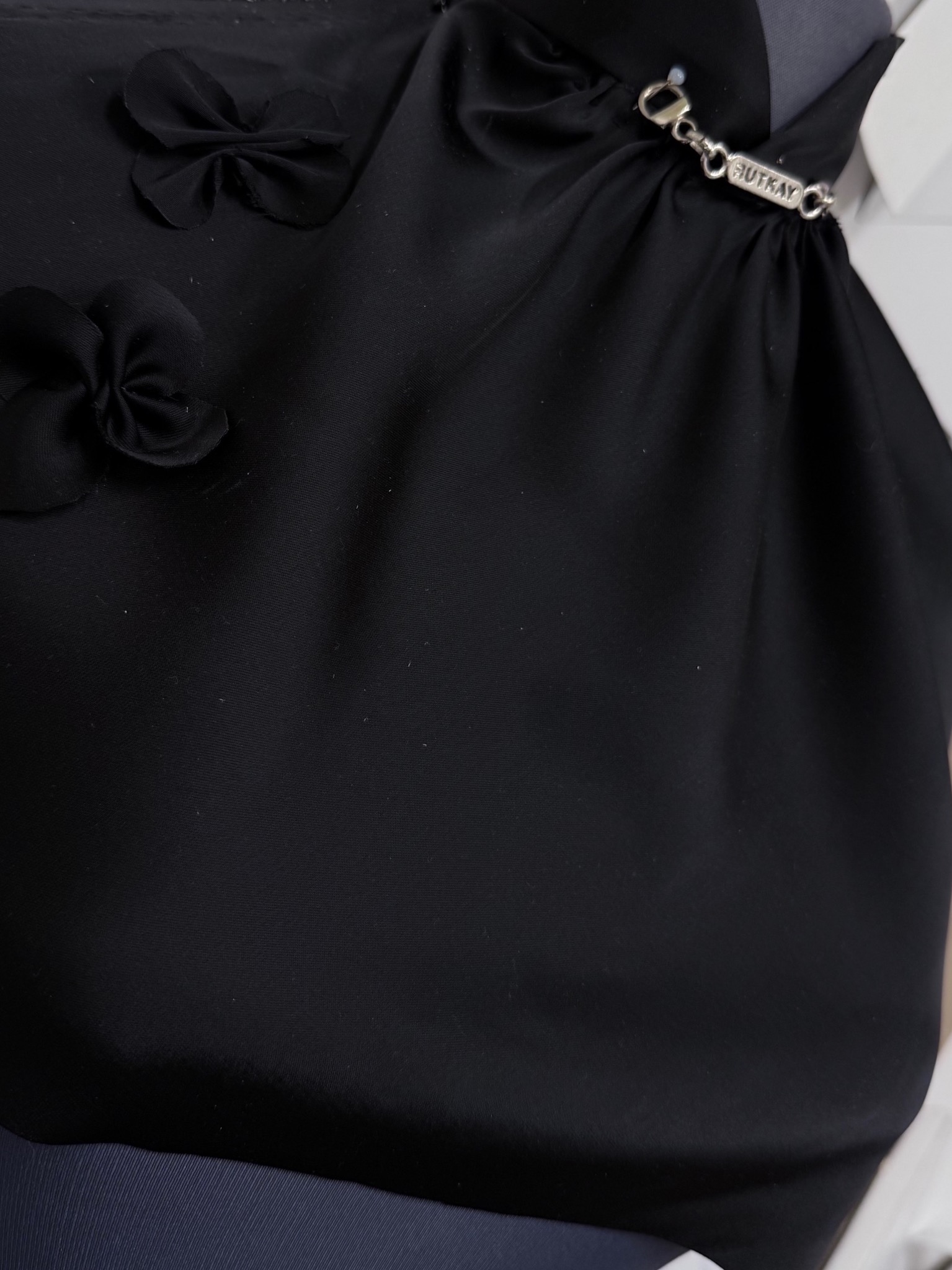 Czarna, elegancka bluzka z ozdobnymi kokardkami i srebrnym łańcuszkiem z napisem 'HUTKAY', widoczna z bliska, prezentująca detale wykończenia.