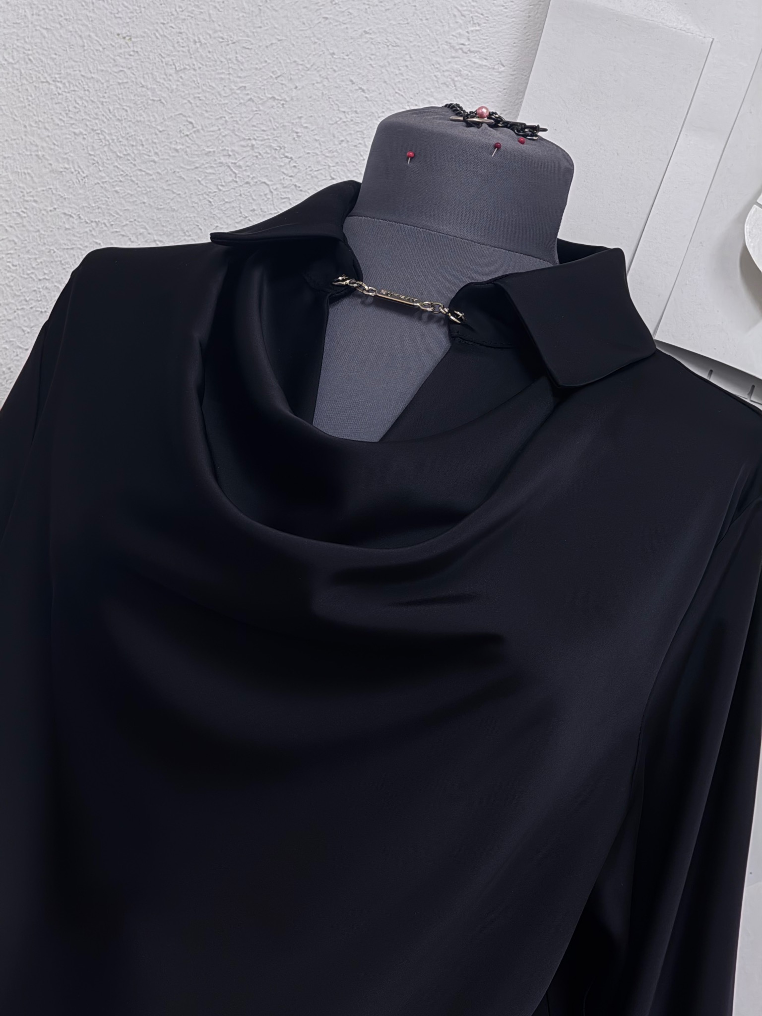 Czarna, satynowa bluzka z drapowanym dekoltem i ozdobnym łańcuszkiem, prezentowana na szarym manekinie krawieckim z przypiętymi szpilkami.