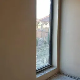 Wnętrze w trakcie wykańczania w Żorach: ściany pokryte świeżym, jasnym tynkiem, widoczne pionowe okno z czarną ramą, przez które widać altanę w ogrodzie. Na podłodze widoczne resztki materiałów...