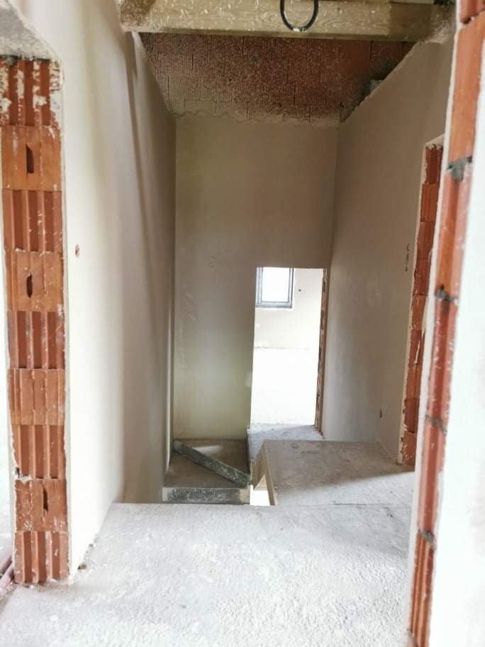 Wnętrze budynku w trakcie prac wykończeniowych, widoczne surowe ściany pokryte świeżo nałożoną warstwą gładzi, fragmenty muru z czerwonej cegły oraz otwór drzwiowy prowadzący do kolejnego...