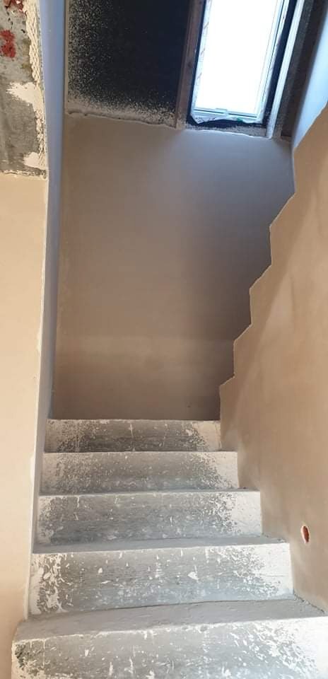 Wnętrze w trakcie prac wykończeniowych: ściana po nałożeniu gładzi w odcieniach beżu, surowe betonowe schody prowadzące na górę i okno dachowe w górnej części kadru.