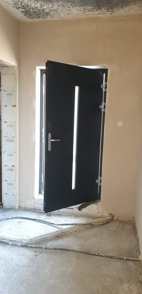 Montaż czarnych drzwi wejściowych z przeszkleniem w surowym wnętrzu po tynkowaniu maszynowym ścian.