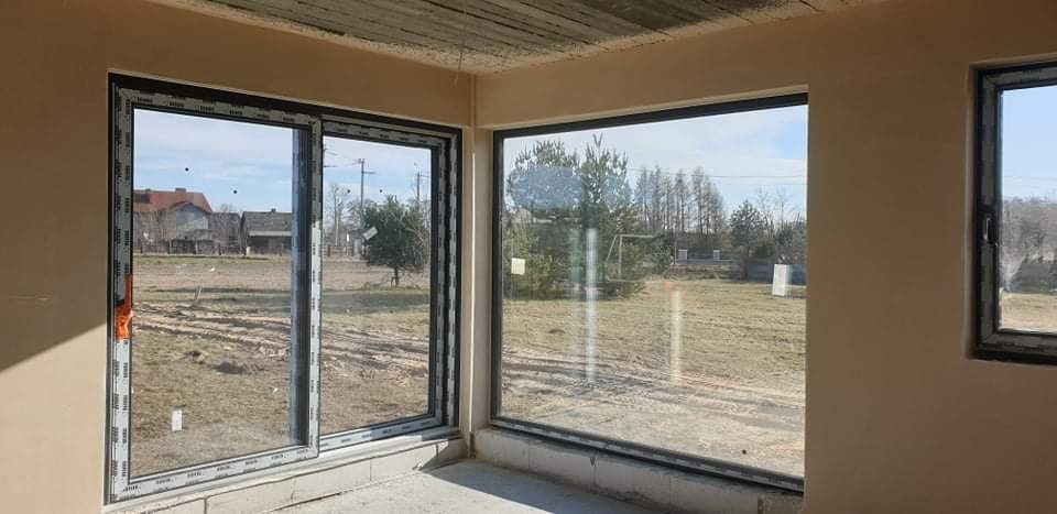 Surowe ściany wewnętrzne z nałożoną warstwą gładzi gipsowej, widoczne okna PCV z widokiem na krajobraz wiejski.