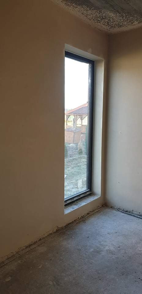 Wnętrze w trakcie wykańczania w Żorach: ściany pokryte świeżym, jasnym tynkiem, widoczne pionowe okno z czarną ramą, przez które widać altanę w ogrodzie. Na podłodze widoczne resztki materiałów...