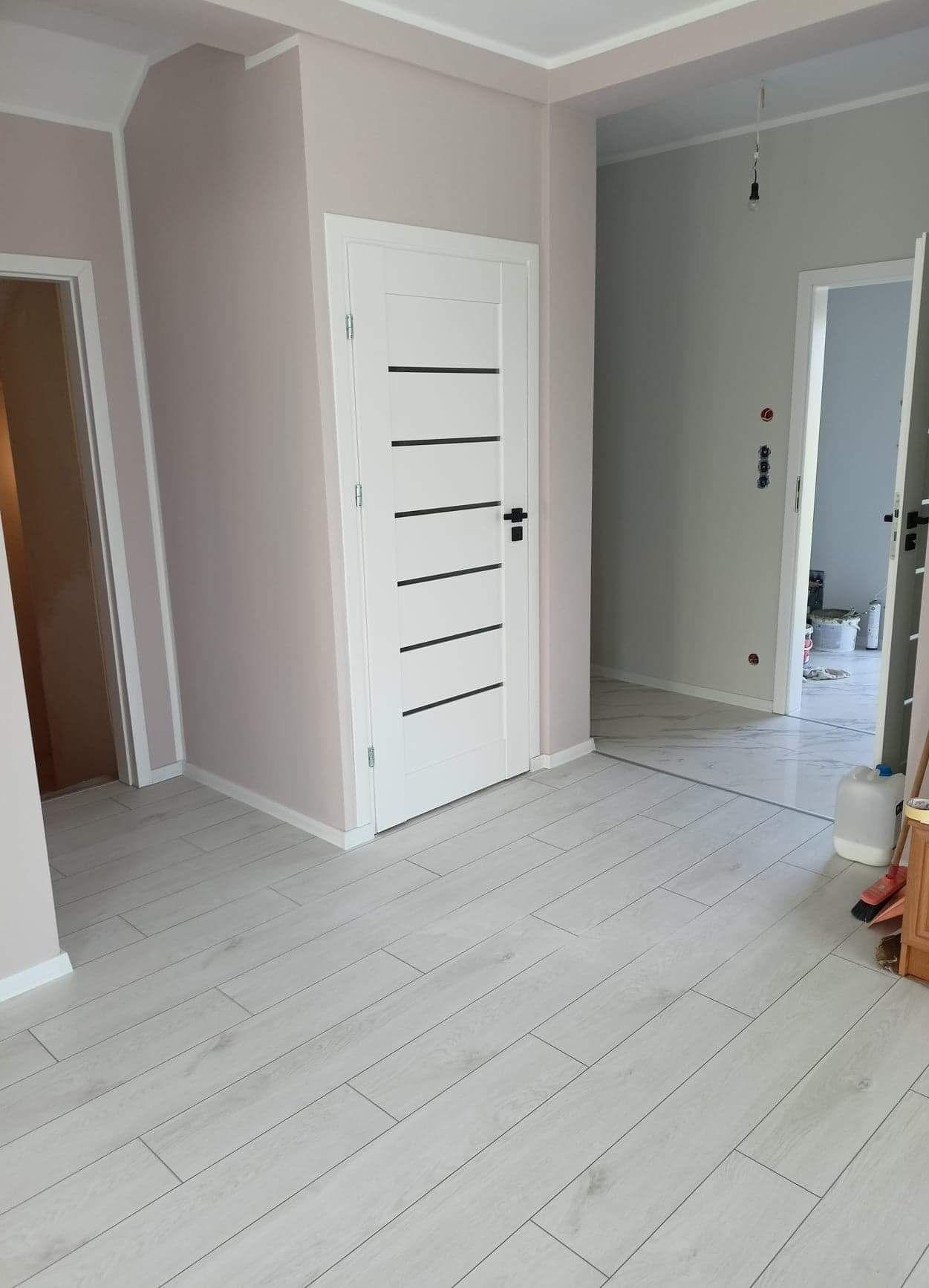 Jasne panele podłogowe ułożone w nowym mieszkaniu, widoczne białe drzwi z czarnymi poziomymi paskami i ściany pomalowane na delikatny róż, w tle widoczne kolejne pomieszczenie w trakcie prac...