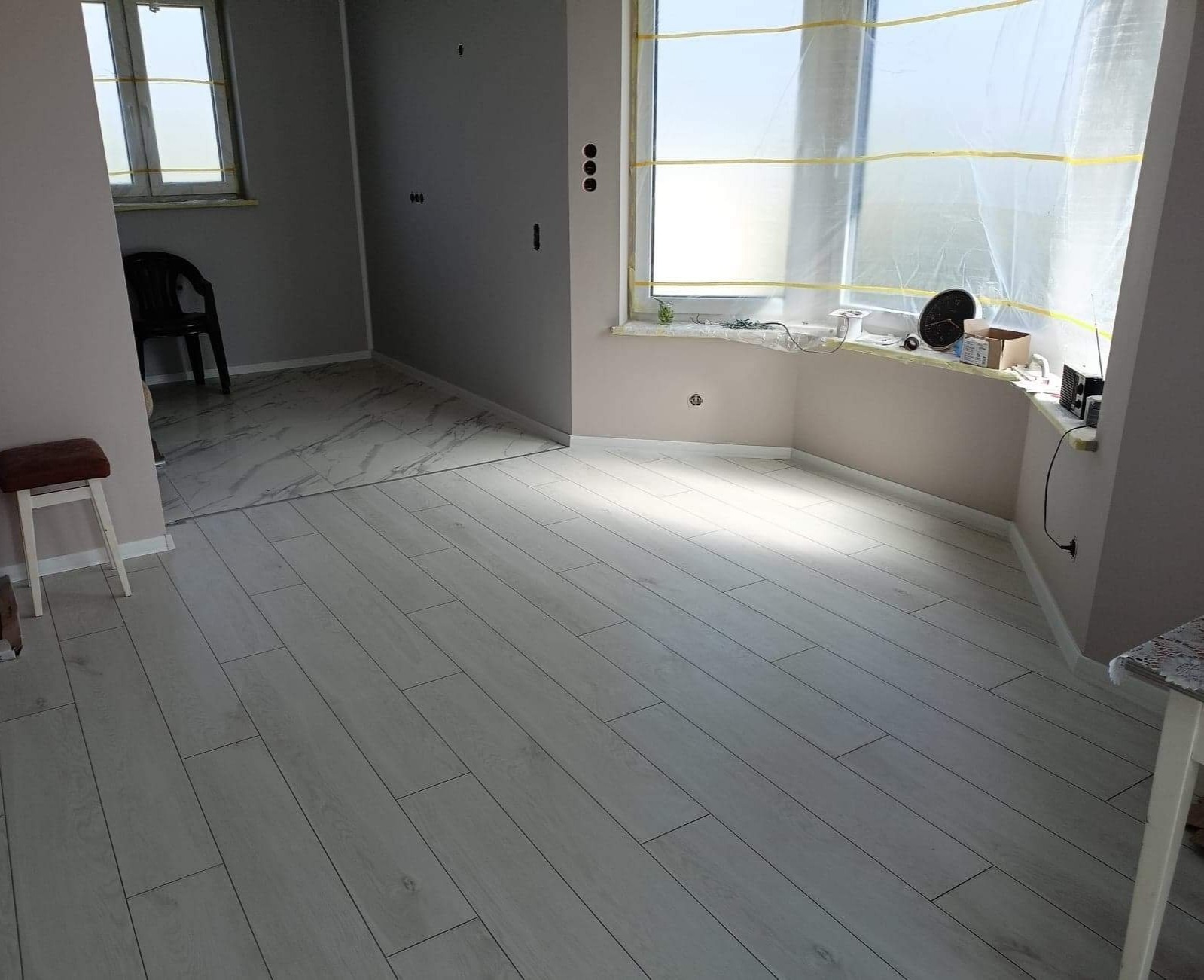 Jasnoszara podłoga panelowa ułożona w pokoju podczas remontu, okno zabezpieczone folią malarską, widoczne gniazdka elektryczne i fragment innego pomieszczenia z marmurową podłogą.