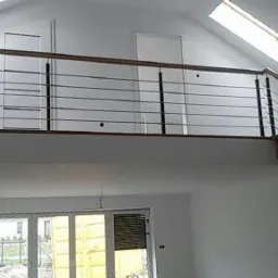 Wnętrze domu z drewnianymi schodami i balustradą ze stali nierdzewnej, widoczne okna dachowe i duże okno na parterze, surowe wykończenie.