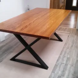 Stół z blatem z jasnego drewna i czarnymi, metalowymi nogami w kształcie litery X, ustawiony na kartonach, w tle białe drzwi i fragment podłogi.