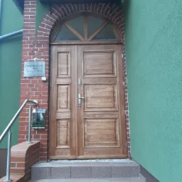 Dwuskrzydłowe, drewniane drzwi zewnętrzne z półokrągłym przeszkleniem nad wejściem, osadzone w ceglanej ramie na tle zielonej ściany budynku, z widocznymi schodami wejściowymi i skrzynką pocztową.