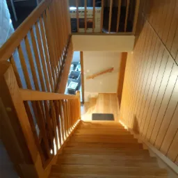 Widok z góry na jasne, drewniane schody z balustradą, prowadzące w dół, z widocznym fragmentem pomieszczenia na dole i panelem ściennym po prawej stronie.