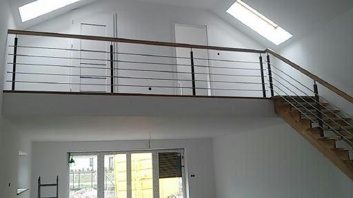 Wnętrze domu z drewnianymi schodami i balustradą ze stali nierdzewnej, widoczne okna dachowe i duże okno na parterze, surowe wykończenie.