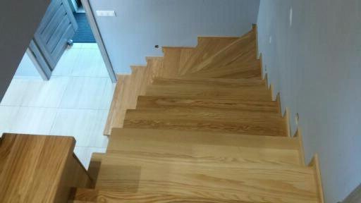Widok z góry na jasne, drewniane schody w domu, z widocznymi słojami drewna i fragmentem drzwi oraz białymi płytkami na podłodze.
