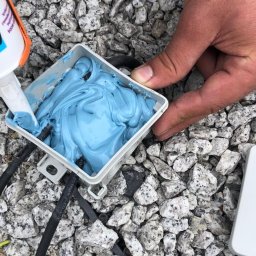 ELECTRO CEL Instalacje elektryczne - Wypełnianie puszki elektrycznej niebieską masą uszczelniającą na tle kamieni, widoczne przewody elektryczne.