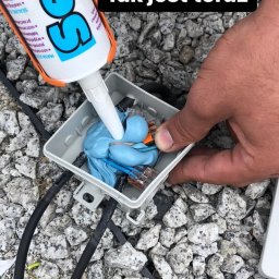 ELECTRO CEL Instalacje elektryczne - Wypełnianie puszki elektrycznej złączkami WAGO i przewodami niebieskim uszczelniaczem silikonowym Sealant, widoczne czarne kable na tle żwirowego podłoża.